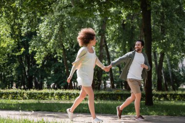 full length of positive young couple holding hands while running together in summer park 