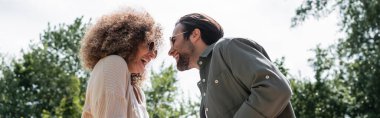 low angle view of cheerful man and happy curly woman in trendy sunglasses looking at each other in park, banner