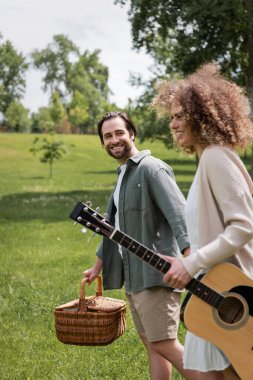 Yeşil parkta sepet taşıyan neşeli adamın yanında akustik gitar tutan mutlu kıvırcık kadın.