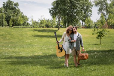 Yeşil parkta yürürken elinde sepet taşıyan adamın yanında akustik gitar tutan mutlu kıvırcık kadın.