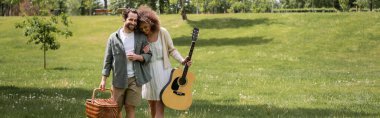 Hasır sepetli adamın yanında akustik gitar tutan mutlu kıvırcık kadın yeşil parkta yürüyor.