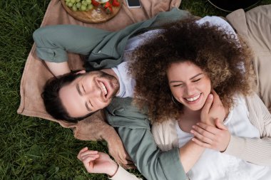 top view of curly woman lying on joyful boyfriend during picnic in summer park
