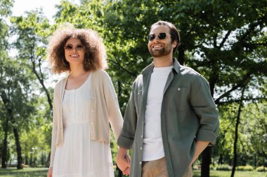 stylish and happy couple in sunglasses holding hands in park