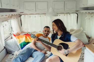 Mutlu ve dövmeli eşcinsel bir adam modern bir minibüste erkek arkadaşının yanında akustik gitar çalıyor. 
