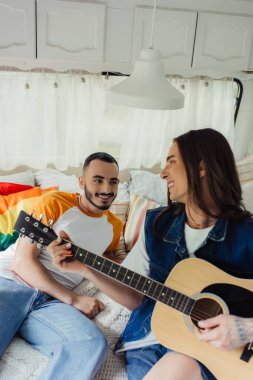 Mutlu ve dövmeli gay erkek arkadaşın yanında akustik gitar çalıyor ve yatakta Igbt bayrağı var. 