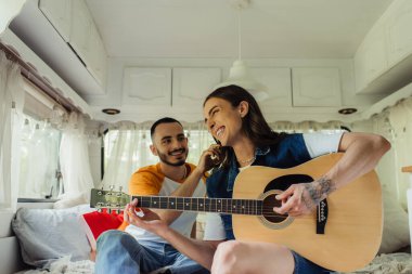Mutlu ve dövmeli eşcinsel bir adam akustik gitar çalıyor. Modern minibüste yatakta gülümseyen bir erkek arkadaşın yanında. 