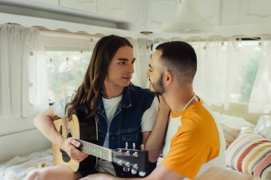 Modern minibüste erkek arkadaşının yanında akustik gitar tutan uzun saçlı eşcinsel bir adam. 