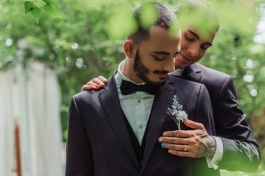 cheerful gay man with wedding ring hugging groom in formal wear with boutonniere