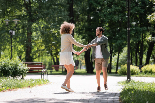 full length of joyful young couple holding hands while walking together in summer park 