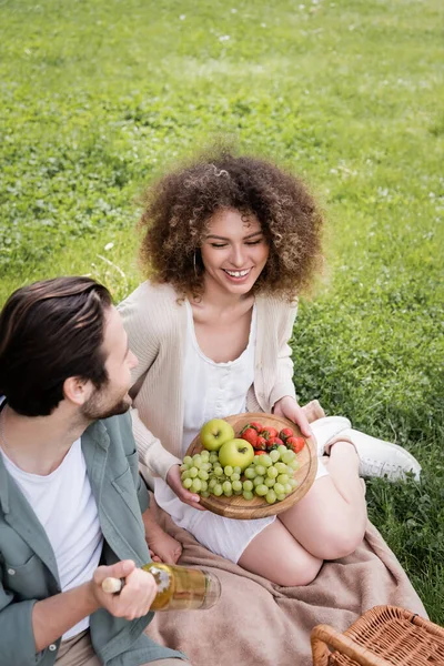 高视角的快乐男人在卷曲的女友旁边拿着一瓶葡萄酒 坐在切菜板和水果旁边 — 图库照片