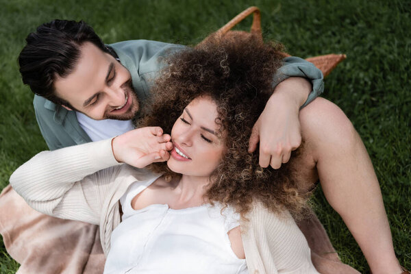 top view of curly woman lying on happy boyfriend during picnic in summer park 