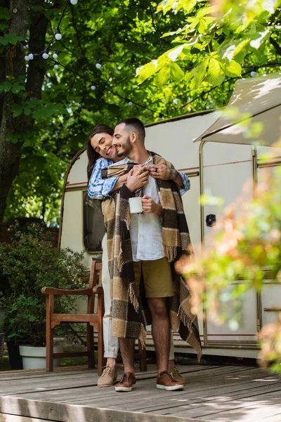 happy tattooed man hugging bearded boyfriend in blanket standing with cup near travel van