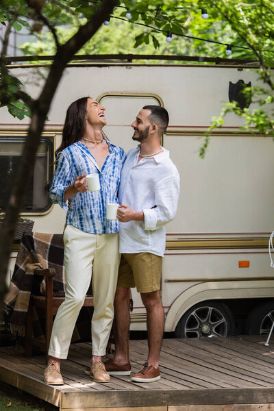 full length of happy gay couple holding cups of coffee while laughing near van in summer