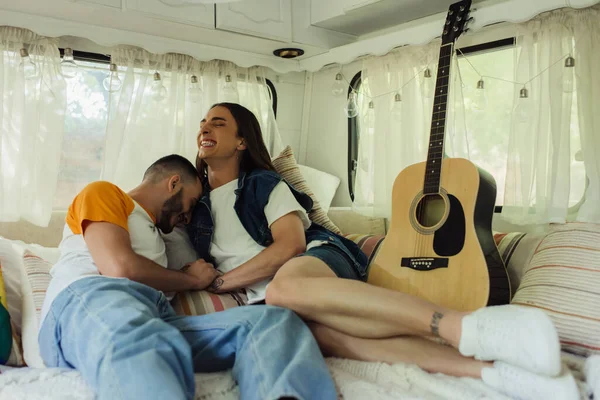 smiling gay man with tattoos laughing with bearded boyfriend on bed in modern van