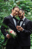 joyful gay couple in suits holding hands near wedding bouquet in green park