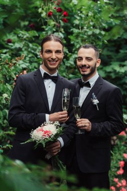 joyful gay couple in suits holding glasses of champagne in green park