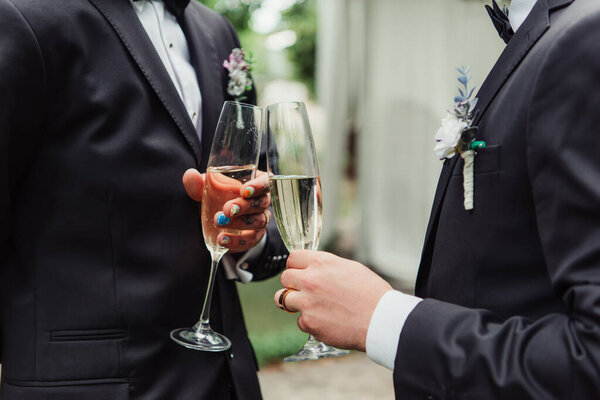 cropped view of gay newlyweds in suits holding glasses with champagne on wedding day 