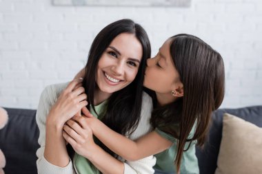 brunette girl with closed eyes kissing cheek of happy babysitter in living room 