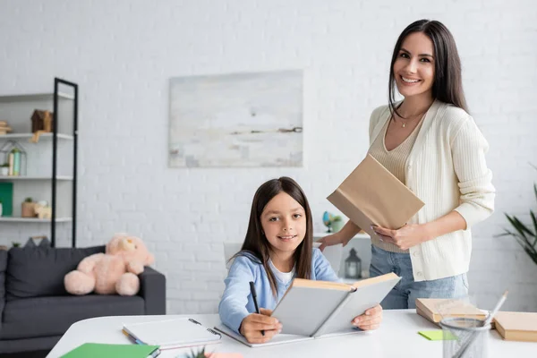 Menina Feliz Fazendo Lição Casa Segurando Livro Perto Babá Sorridente — Fotografia de Stock