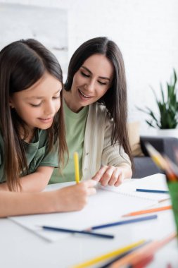 cheerful babysitter looking at paper while girl drawing with yellow pencil 