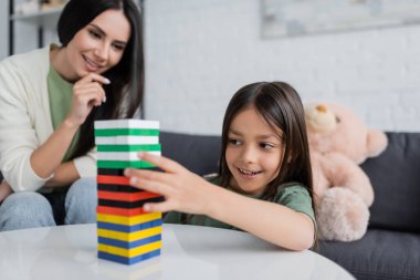 Olumlu bebek bakıcısı oturma odasında memnun çocukla ahşap kule oyunu oynuyor.