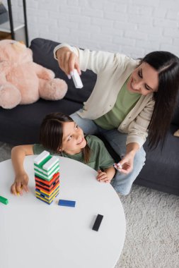 Oturma odasında mutlu bir kızla ahşap kule oyunu oynayan pozitif bakıcının yüksek açılı görüntüsü.