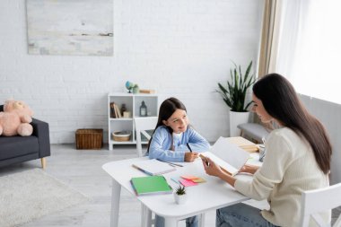 nanny reading book near smiling girl doing homework in modern living room