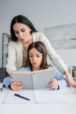 brunette nanny helping girl reading book while doing homework