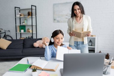 smiling girl holding copybook in front of laptop near nanny with book on background