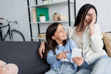 laughing nanny obscuring face with hand near girl pointing at smartphone on couch at home