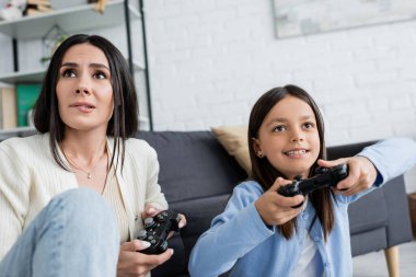 KYIV, UKRAINE - MAY 23, 2022: worried babysitter biting lip while gaming with cheerful girl at home