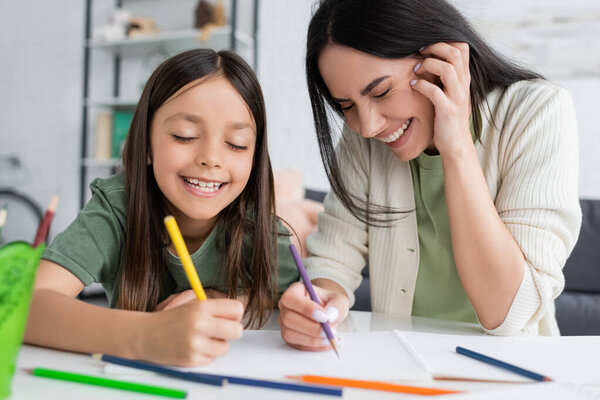 happy babysitter with closed eyes laughing while cheerful girl drawing on paper