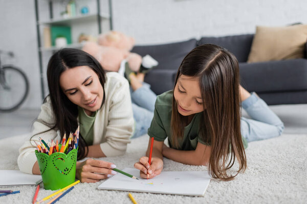 smiling nanny and girl drawing on paper while lying on carpet in living room 