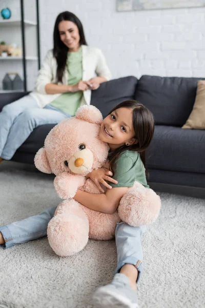 Happy Girl Sitting Carpet Hugging Teddy Bear Blurred Babysitter Living — Zdjęcie stockowe