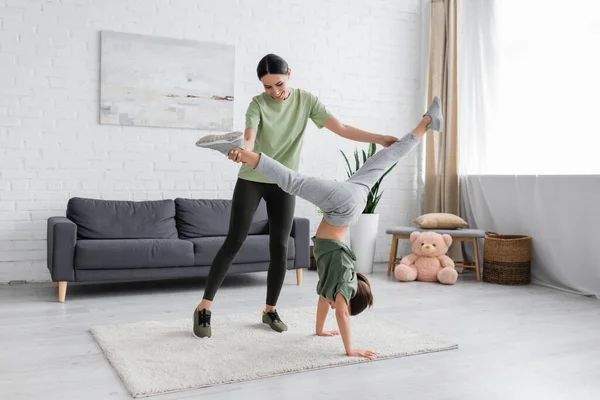 Smiling Babysitter Supporting Girl Doing Handstand Living Room — Zdjęcie stockowe