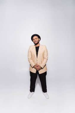 full length of stylish indian hip hop performer in fedora hat and eyeglasses standing on grey