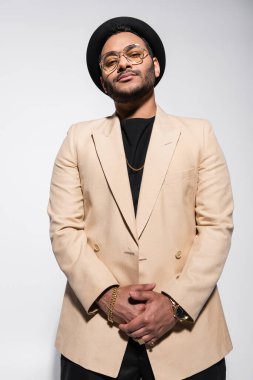 low angle view of stylish indian hip hop performer in fedora hat and eyeglasses looking at camera isolated on grey