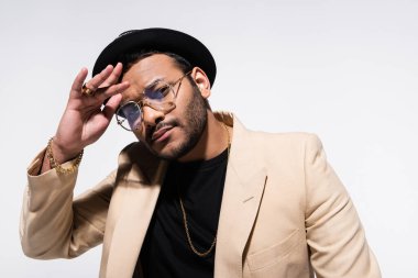 Fötr şapkalı ve gözlüklü şık Hintli hip hop sanatçısı gri renkli kameraya bakarken el kol hareketi yapıyor.