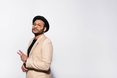 stylish indian rapper in fedora hat and eyeglasses showing peace sign isolated on grey