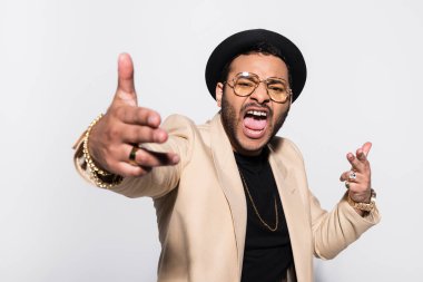 emotional indian hip hop performer in fedora hat and eyeglasses screaming while gesturing isolated on grey