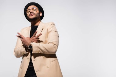 low angle view of indian hip hop performer in fedora hat and eyeglasses gesturing isolated on grey