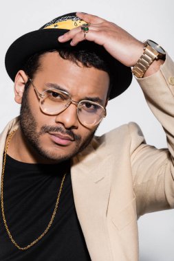 portrait of eastern hip hop performer in eyeglasses adjusting fedora hat isolated on grey