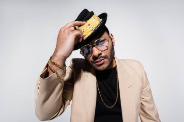 eastern hip hop performer in eyeglasses holding fedora hat and looking at camera isolated on grey