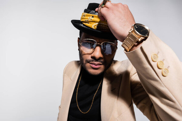 eastern hip hop performer with golden wristwatch holding fedora hat and looking at camera isolated on grey