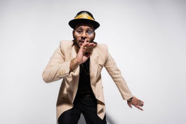 eastern hip hop performer in fedora hat and eyeglasses showing hush sign on grey