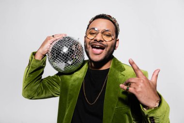 excited indian hip hop performer in green velvet blazer and crown holding disco ball isolated on grey