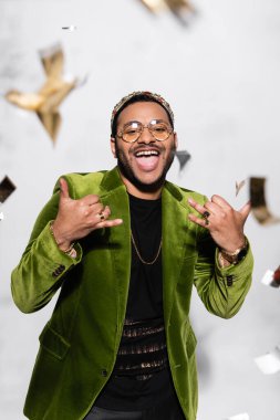 excited indian hip hop performer in green velvet blazer and crown gesturing near falling confetti on grey