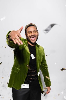 excited indian hip hop performer in green velvet blazer and crown singing near falling confetti on grey