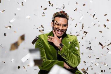 cheerful indian hip hop performer in green velvet blazer and crown near falling confetti on grey