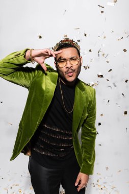 indian hip hop performer in green velvet blazer and crown pouting lips near falling confetti on grey